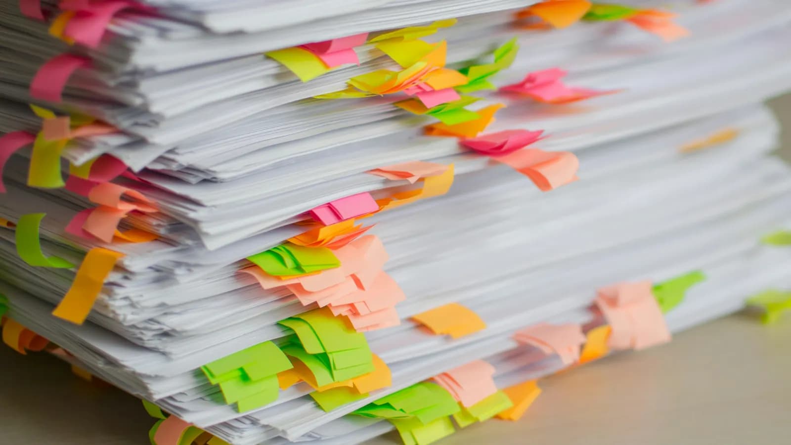 A tall stack of printed papers on a desk, its edges flagged with pink, orange, yellow and green sticky tabs.