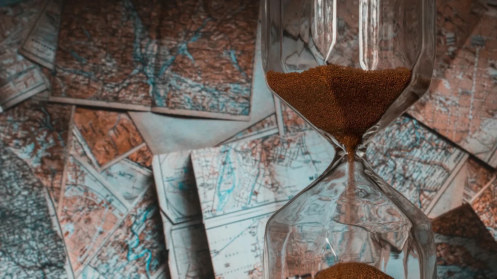 A glass hourglass with brown sand running through it, standing on a table covered in old paper maps.