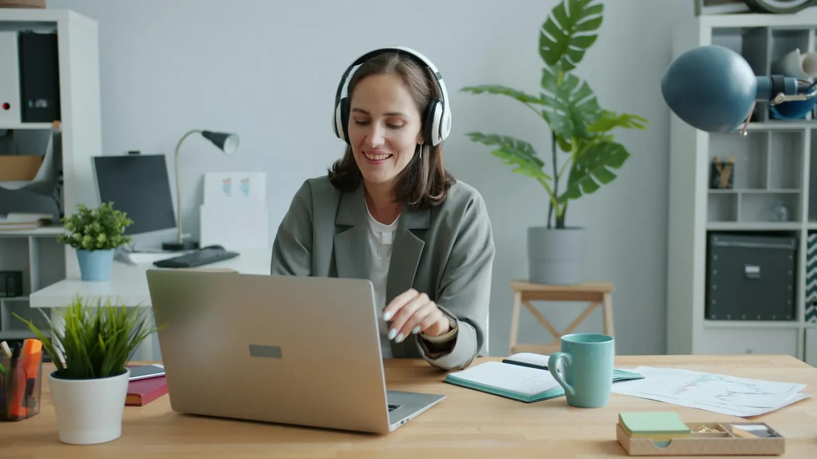 A smiling woman wearing headphones works on a laptop at a desk in a bright home office with plants and shelves.