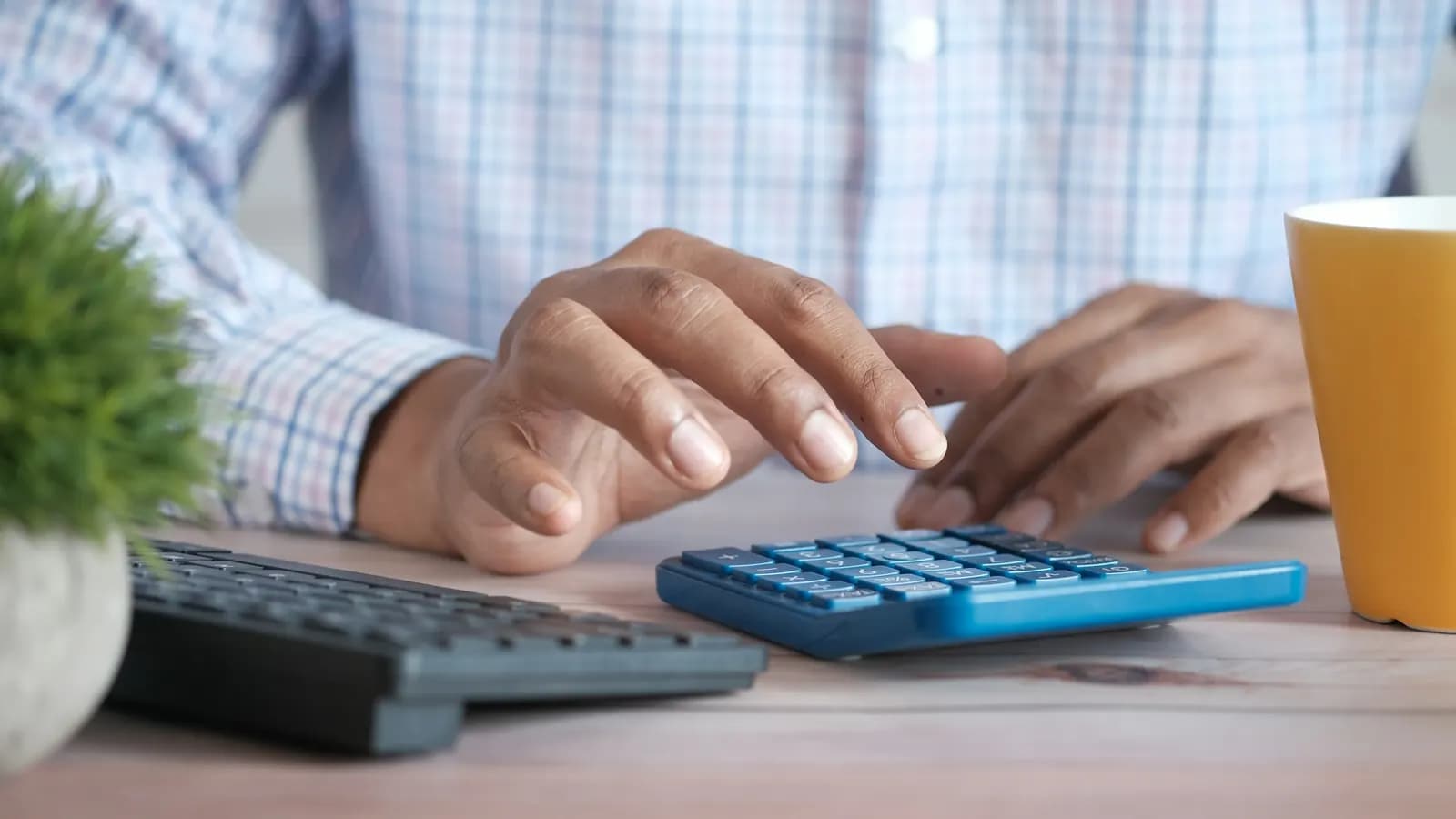 Close-up of a person's hands using a blue calculator on a desk beside a keyboard and a yellow mug.