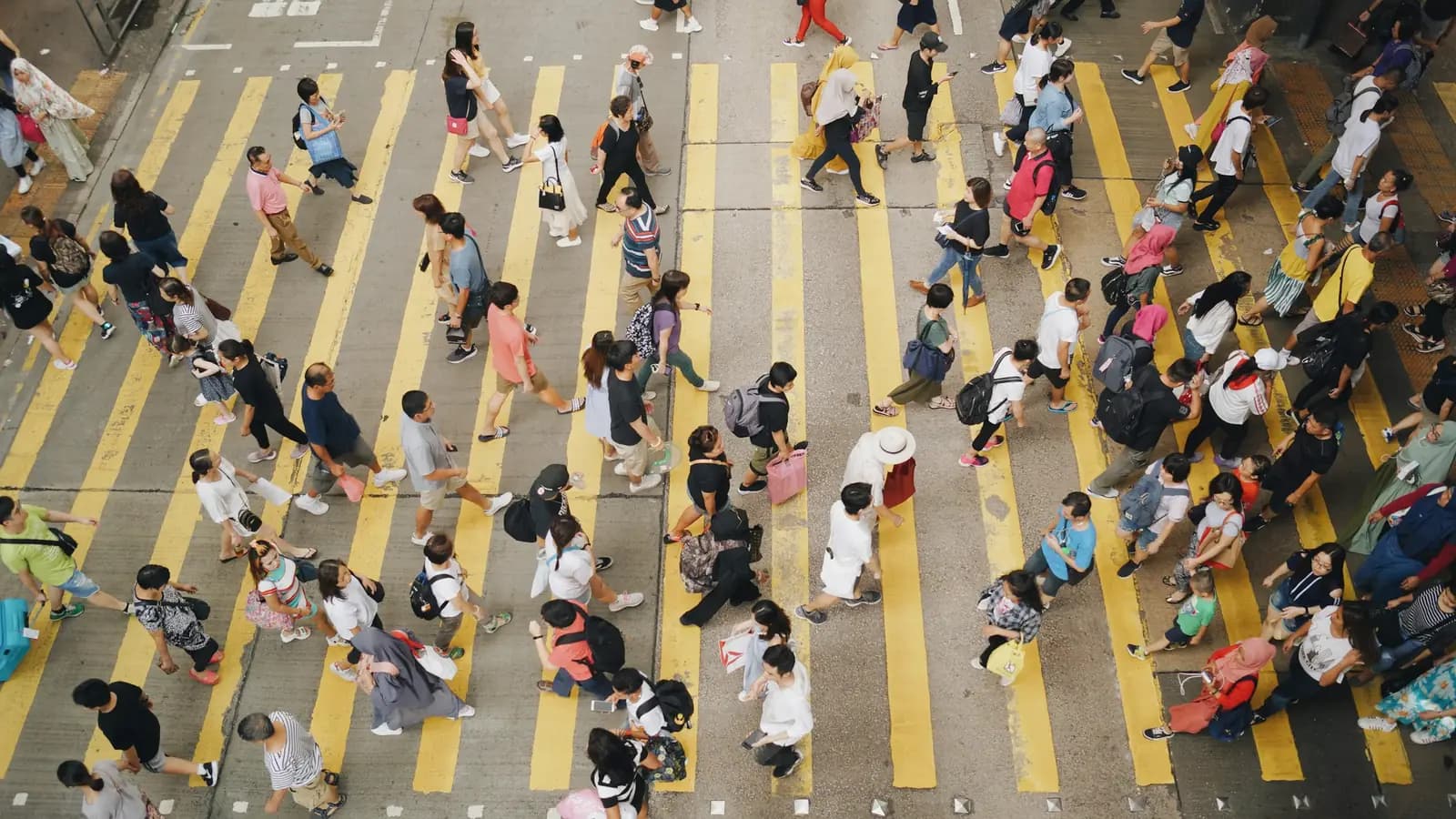 Seen from above, a crowd of people crosses a wide pedestrian crossing painted with yellow stripes on a street in Mong Kok, Hong Kong.