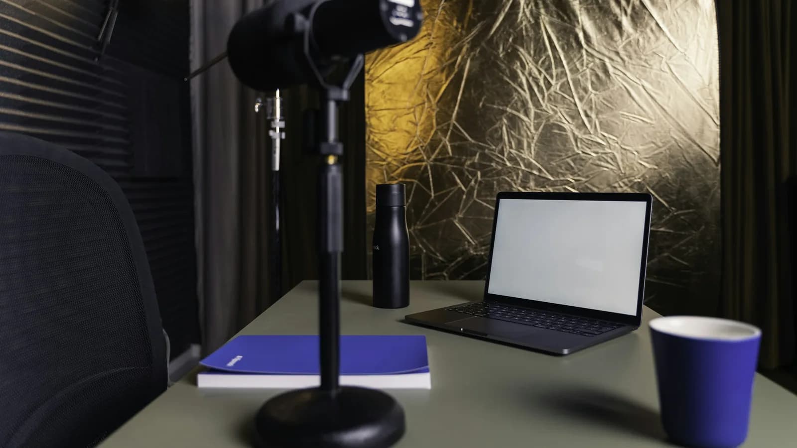 A microphone on a desk stand in front of an open laptop, with a blue notebook and a blue cup on the desk.