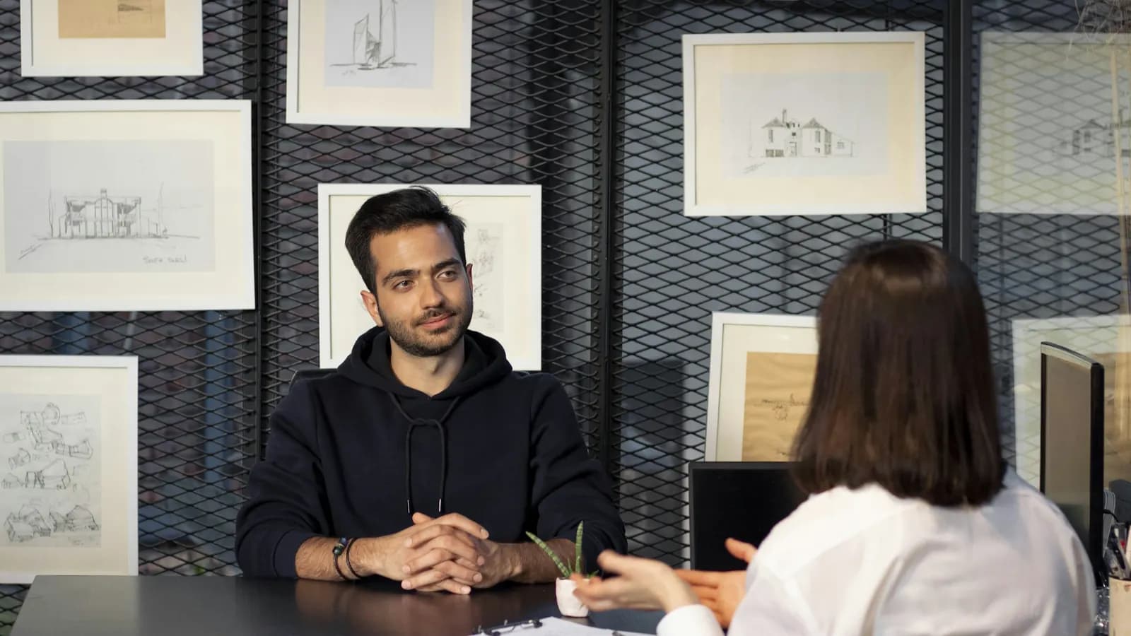 A man sits with folded hands across a desk from a woman, seen from behind, in an office with framed sketches on the wall.