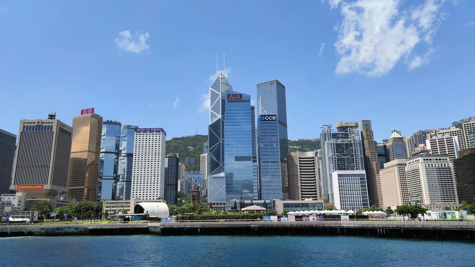The Central District waterfront on Hong Kong Island, with office towers behind the promenade and a boat passing on the harbour.