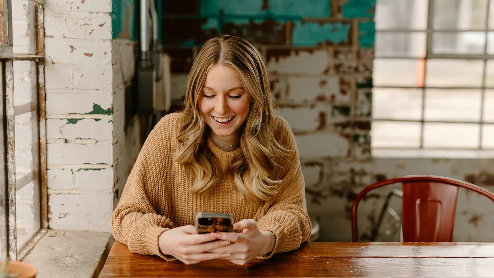 A smiling woman in a knit sweater sits at a wooden table looking at her phone.
