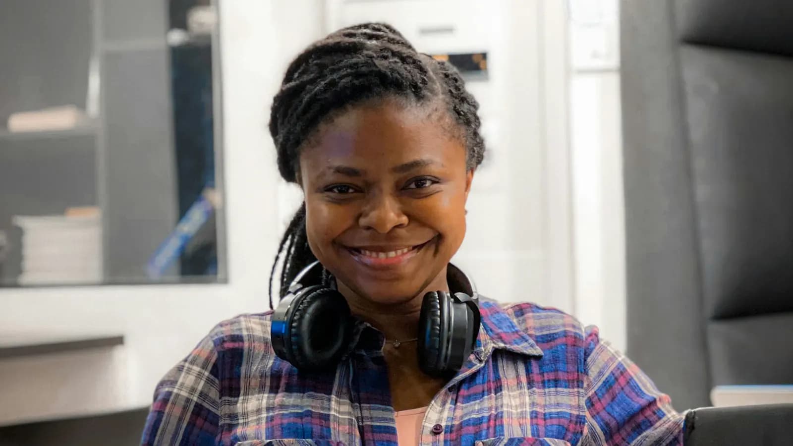 A smiling woman with headphones around her neck sits at a desk beside an open laptop.