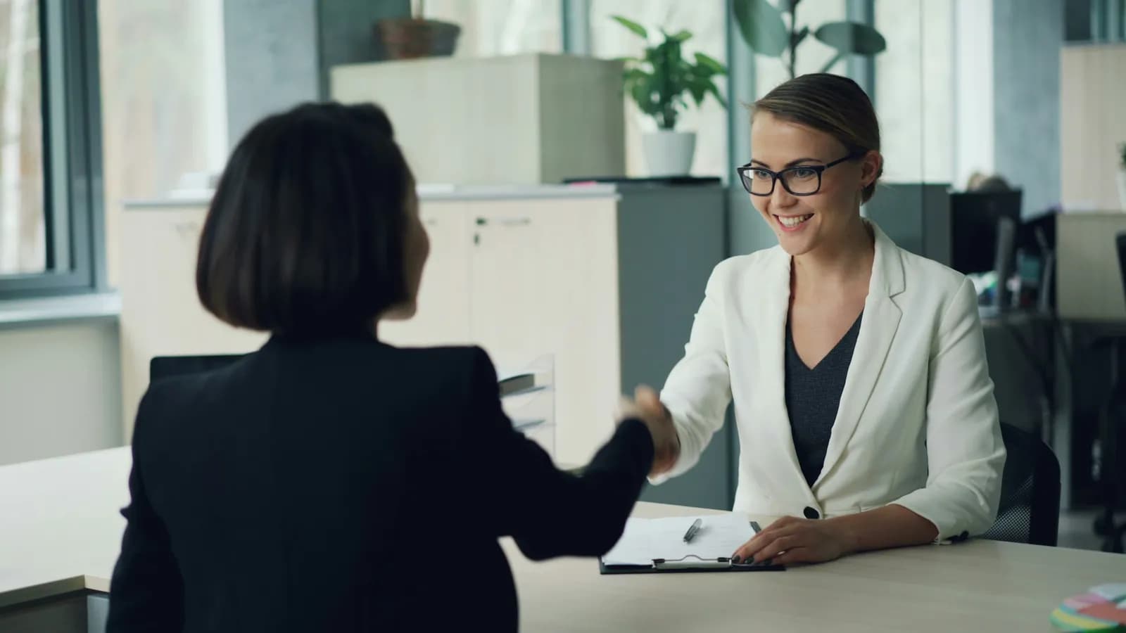 A woman in glasses and a white blazer smiles and shakes hands across a desk with a woman seen from behind, in a bright office.