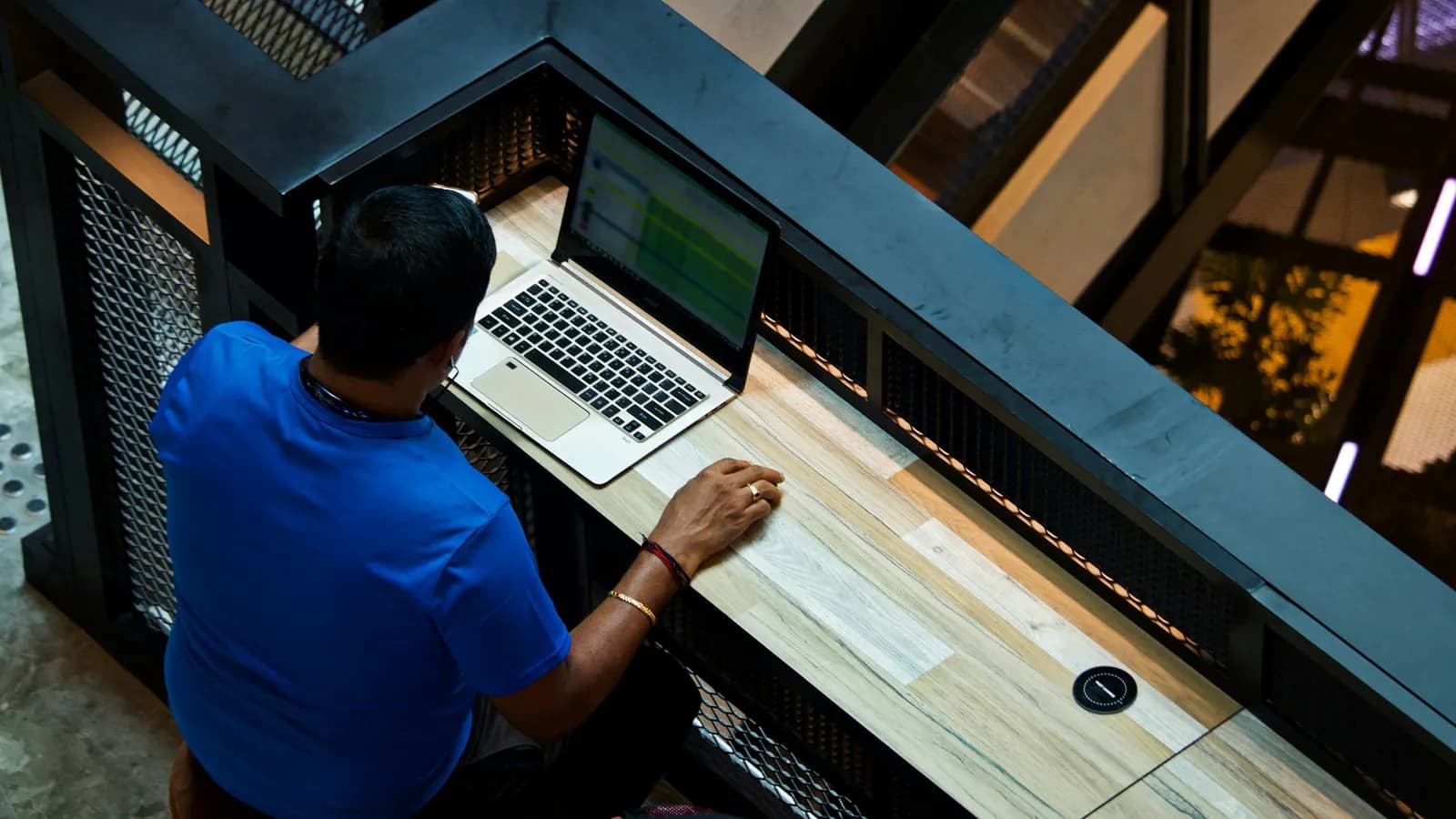 Seen from above, a man in a blue T-shirt sits at a wooden counter with an open laptop, his hand resting beside it.