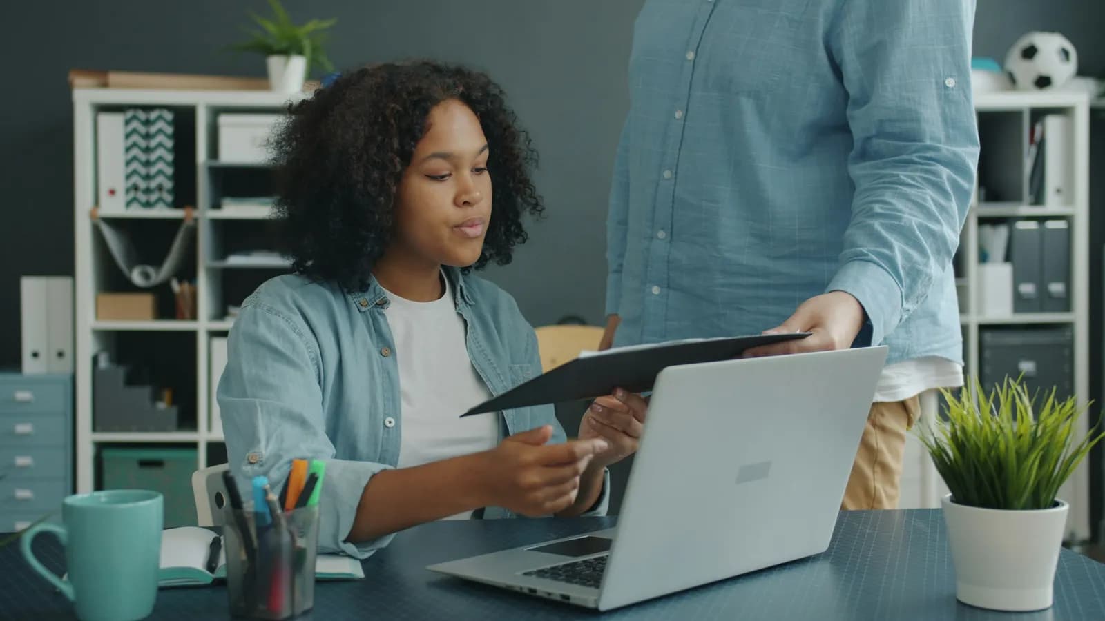 A woman at an office desk takes a folder from a colleague standing beside her, with an open laptop in front of her.