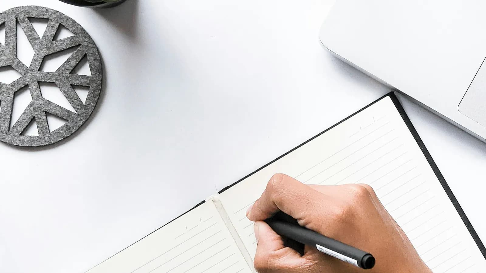 Overhead view of a hand writing in an open notebook on a white desk, next to a laptop and a felt coaster.