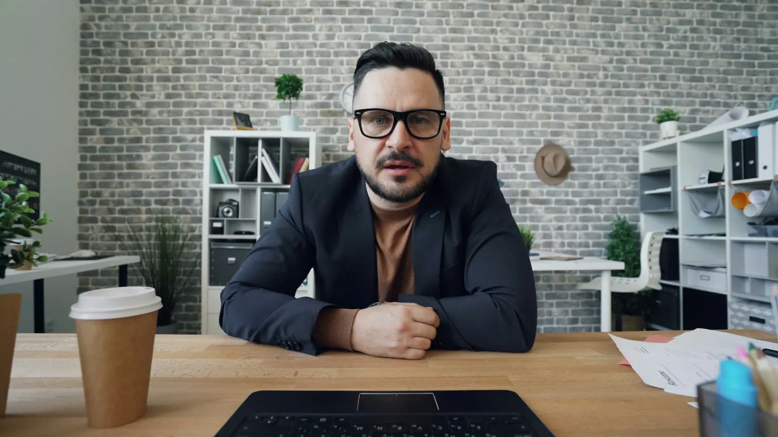 A man in glasses and a blazer sits at a desk and looks straight into his laptop's camera, with a paper coffee cup beside him.