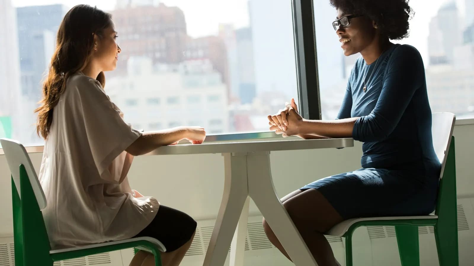 Two women talk across a white table by a large window overlooking city buildings.